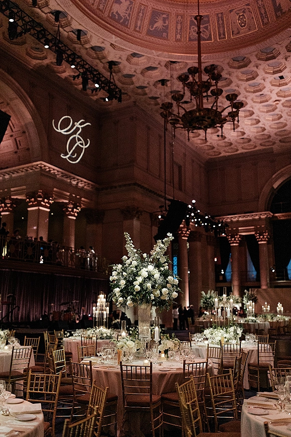 Reception tablescape with a tall floral centerpiece of white roses and greenery, candles, and gold chiavari chairs in a grand ballroom