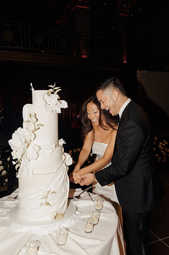 Wedding cake cutting as bride in strapless dress and groom in black tux slice a white tiered cake with sugar flowers in dim reception light