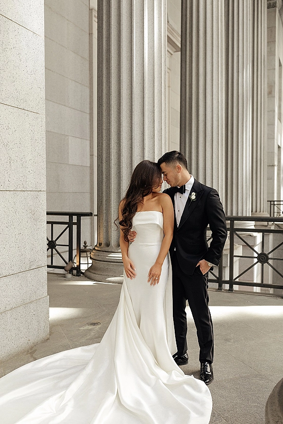Couple portrait of bride and groom embrace in a forehead touch pose, her strapless gown with long train by stone columns outside a building