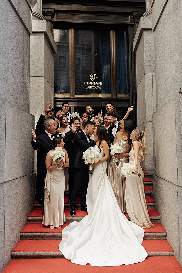 Wedding party photo of bride and groom kiss as friends cheer on red carpet stairs by a stone building entrance with glass doors