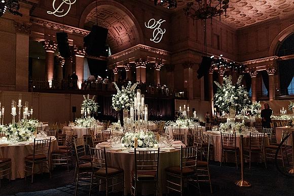 Reception tablescape with tall glass taper candles and white-green floral centerpieces on round tables in a grand ballroom with arched ceilings