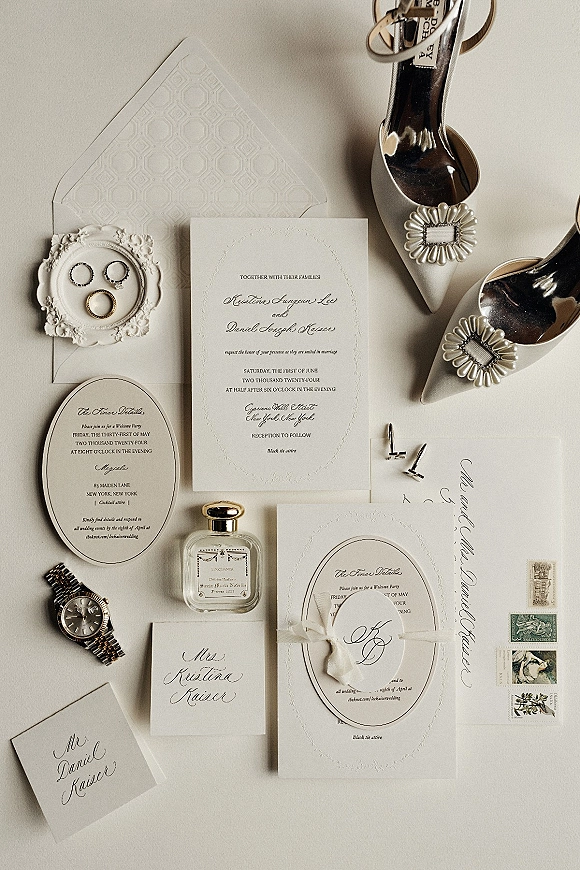 Invitation suite wedding invitation flat lay with embossed cards, calligraphy envelope, wax seal, rings, shoes, and cufflinks on neutral tabletop