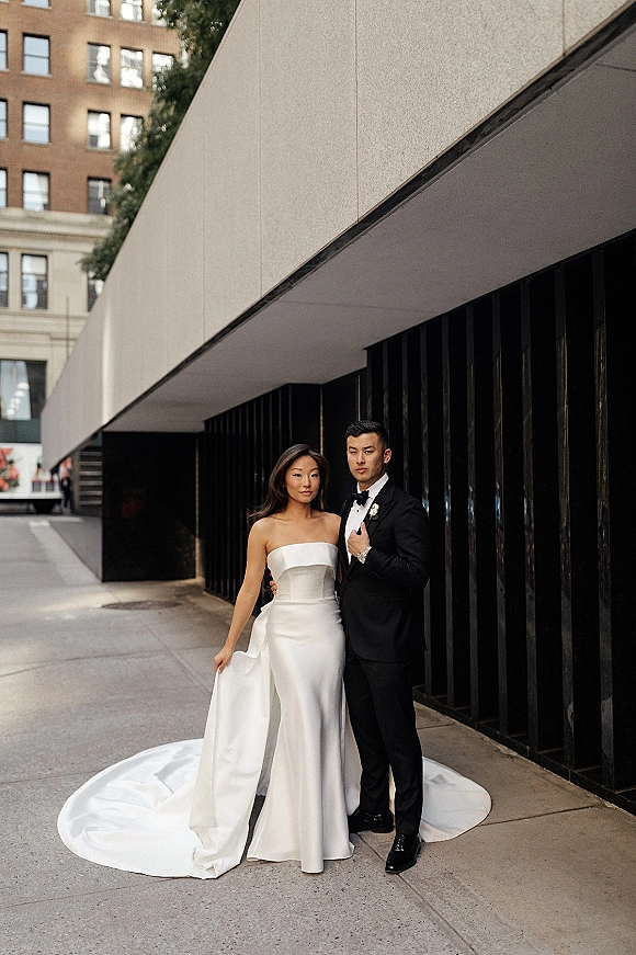 Couple portrait of bride in a strapless wedding dress holding her long train beside groom in black tuxedo on a city sidewalk by slat wall