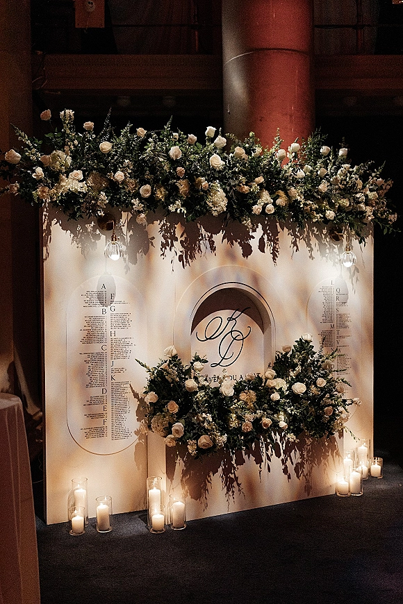 Seating chart display with arched wedding seating chart cards, white florals and greenery, candlelight and pendant lights in an industrial venue