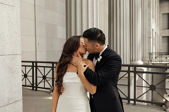 Wedding kiss portrait of bride and groom kissing as he cups her face, with strapless gown and black tuxedo on a stone-column balcony