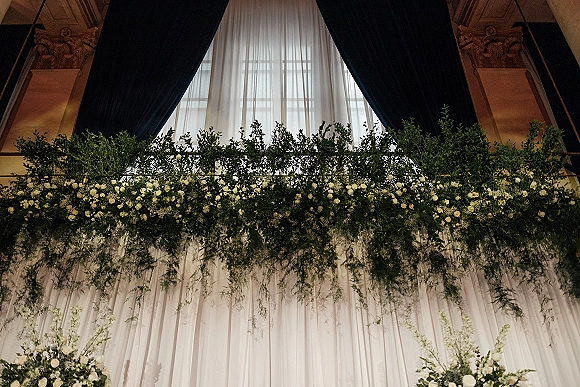 Wedding floral installation with hanging greenery wedding decor, white roses and baby’s breath over sheer draping in a grand ballroom by a tall window