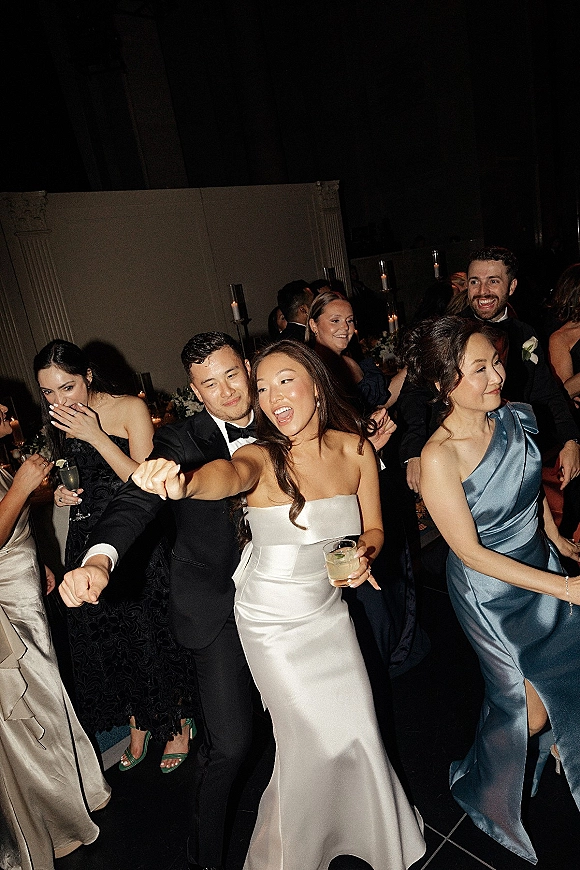 Wedding reception dance as bride in a strapless satin gown and groom in a black tuxedo sway with guests on a dim dance floor, candlelit tables behind