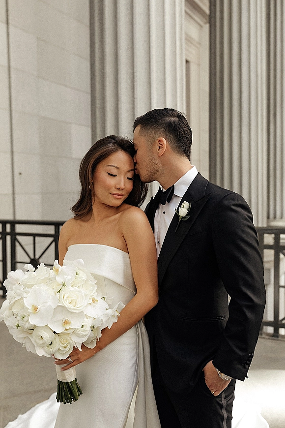 Couple portrait of groom kissing bride’s forehead as she holds a white rose bouquet, standing by stone columns and railing outdoors
