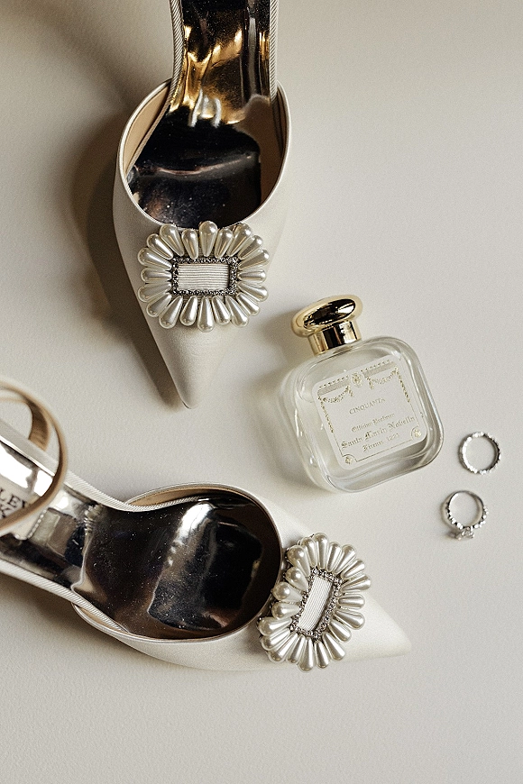 Bridal shoes flatlay with white satin wedding heels, pearl and rhinestone buckles, perfume bottle, rings, and ribbon on neutral surface
