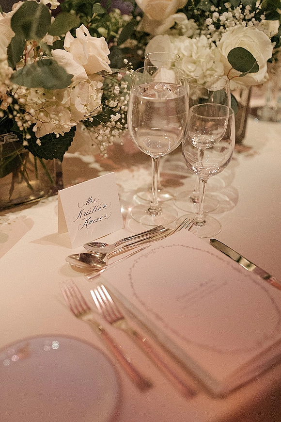 Reception tablescape with a wedding place setting, white rose and hydrangea centerpiece, and soft ambient lighting in the background