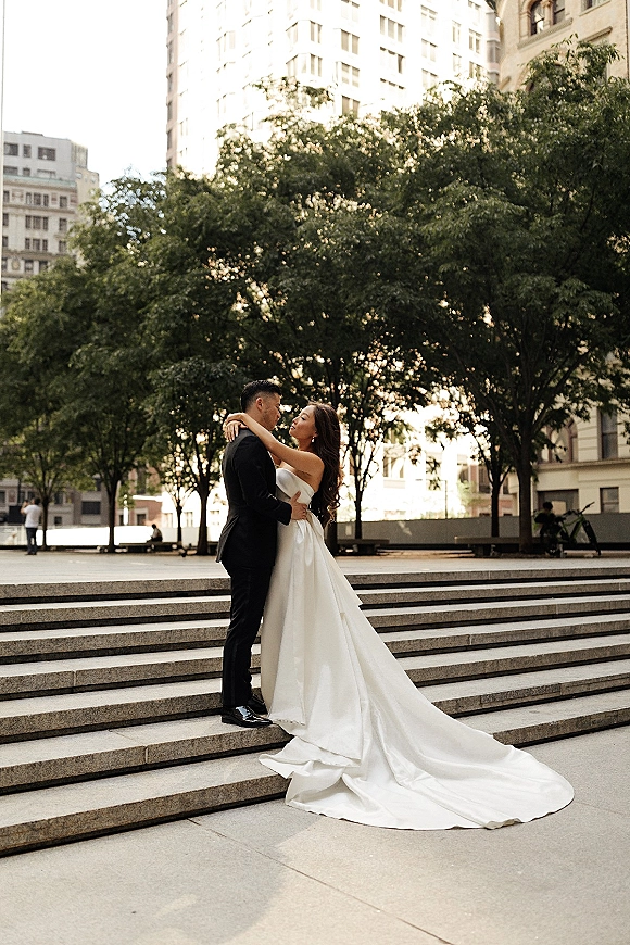 Couple portrait of bride and groom embrace on stone steps, her strapless gown’s long train spread as city buildings and trees frame them