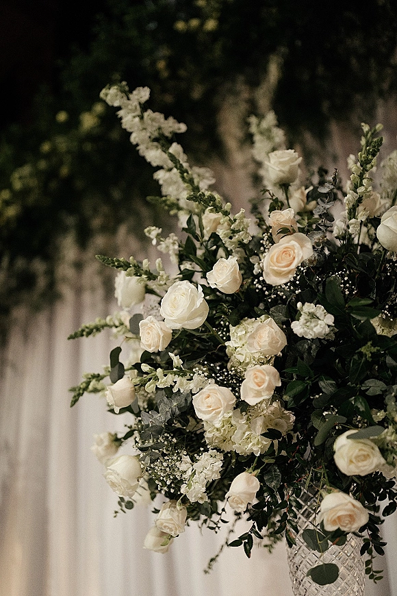 Wedding floral arrangement, white rose centerpiece in a cut glass vase with hydrangea and greenery against white drapery and hanging greens