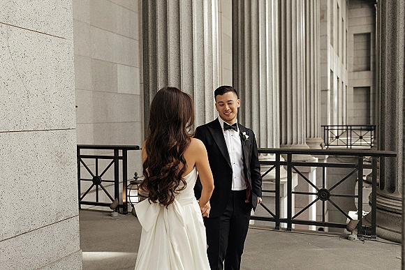 First look moment as bride in strapless wedding dress meets groom in black tuxedo with boutonniere on a balcony by stone columns