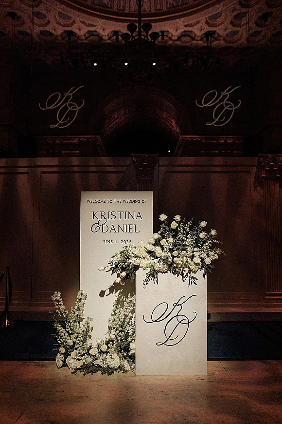 Wedding welcome sign with white rose and greenery florals at the base in an ornate indoor venue with balcony railing and chandelier
