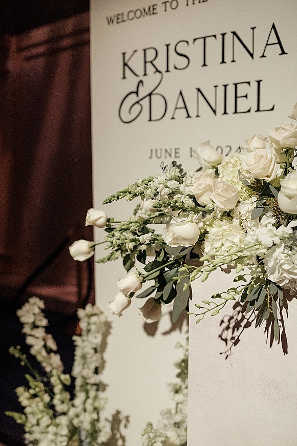 Wedding welcome sign with flowers on a foam board, styled with white roses, hydrangea, and greenery against a wood door in soft shadow light