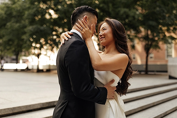 Couple portrait of bride and groom embrace on outdoor steps, bride’s ringed hand on his face beside trees and a building facade