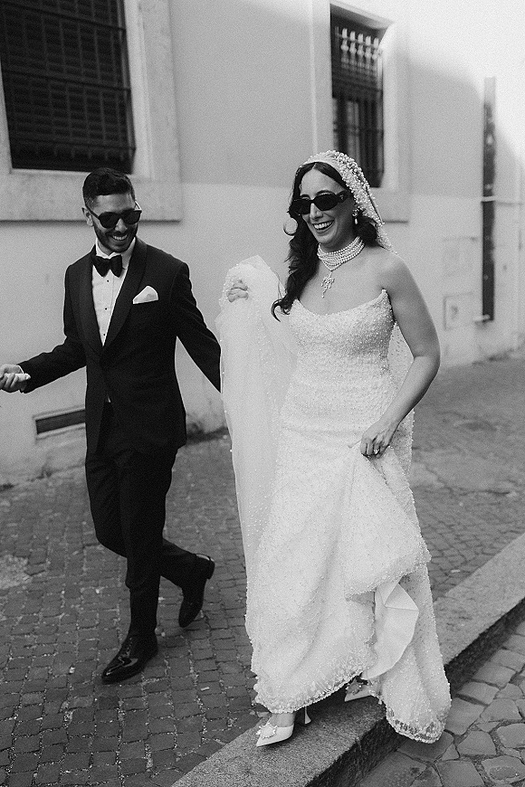 Couple portrait in a black and white wedding photo, bride and groom walking with sunglasses on a cobblestone street by building windows