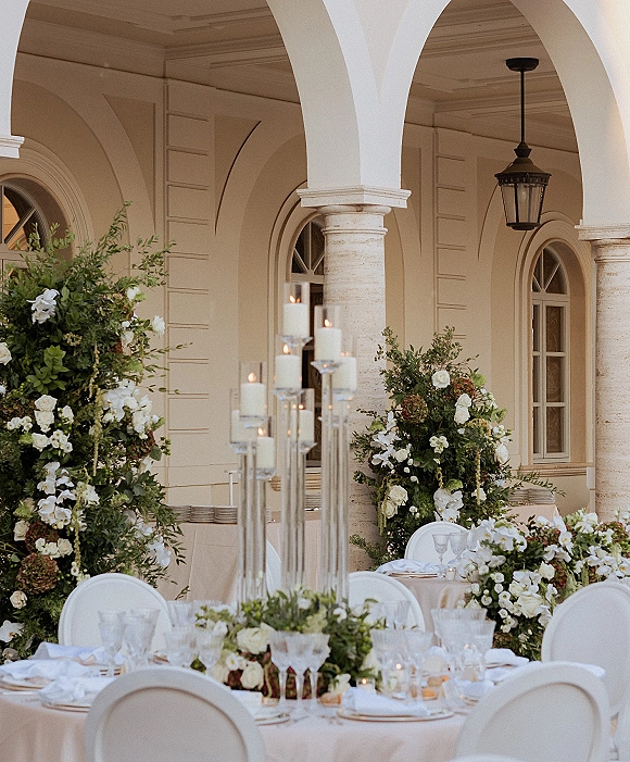 Reception tablescape with candle centerpiece wedding tall glass candlesticks, white florals and greenery garland under an arched colonnade with lanterns