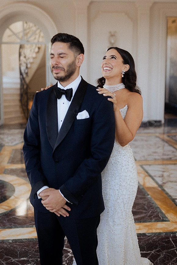 First look moment as bride taps groom’s shoulder in a marble hallway with arched doorway, her pearl choker gleaming beside his tuxedo