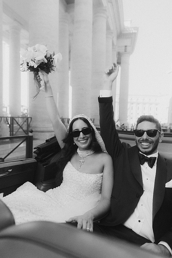 Couple portrait of newlyweds in convertible, both in sunglasses, bride lifting bouquet beside groom in tux, city columns behind