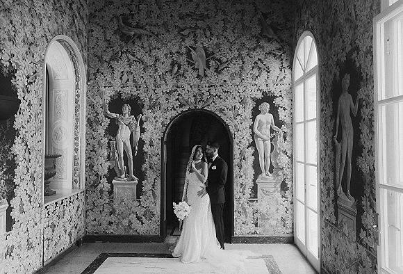 Couple portrait in a black and white wedding portrait style, bride and groom embrace in an arched doorway with window light and statues behind