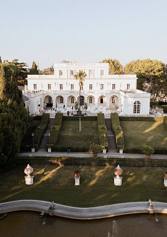 Outdoor reception setup with white round tables and floral arrangements under string lights beside a white villa terrace and fountain