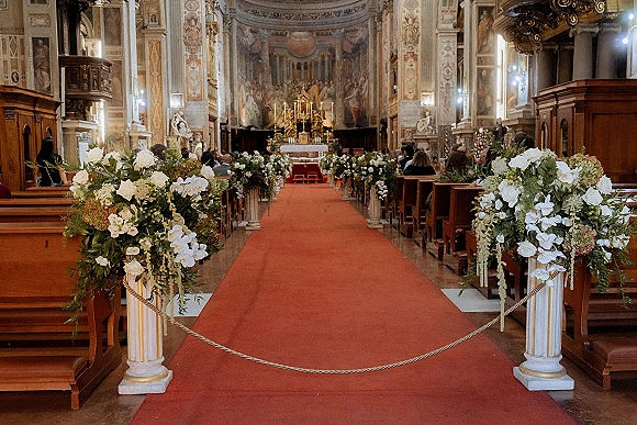 Church ceremony aisle lined with wedding aisle florals of white roses, orchids and hydrangeas on pedestals, red runner to candlelit altar
