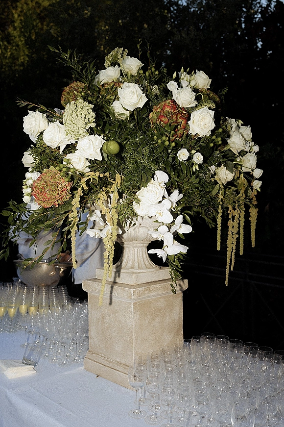 Wedding floral arrangement in a stone urn wedding centerpiece with white roses, hydrangea and greenery on a bar table with champagne flutes at night outdoors