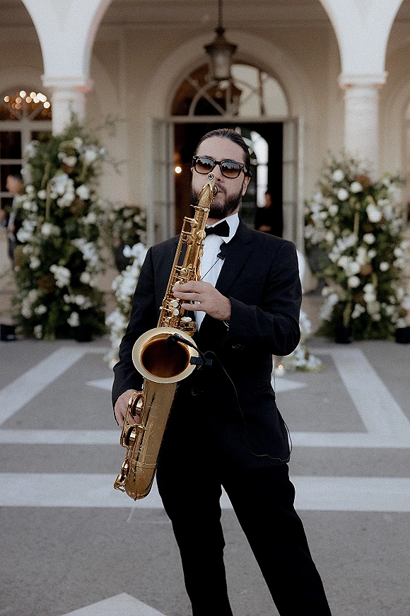 Wedding saxophonist providing saxophonist wedding entertainment in a tuxedo and sunglasses, playing by an arched entrance with lanterns and florals