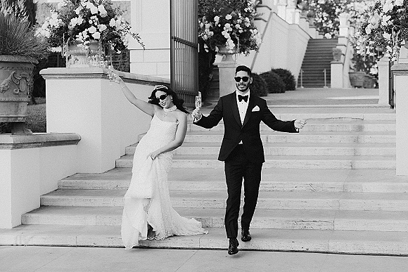 Wedding couple portrait of bride and groom in sunglasses toasting with champagne flutes on stone steps with large floral urns behind