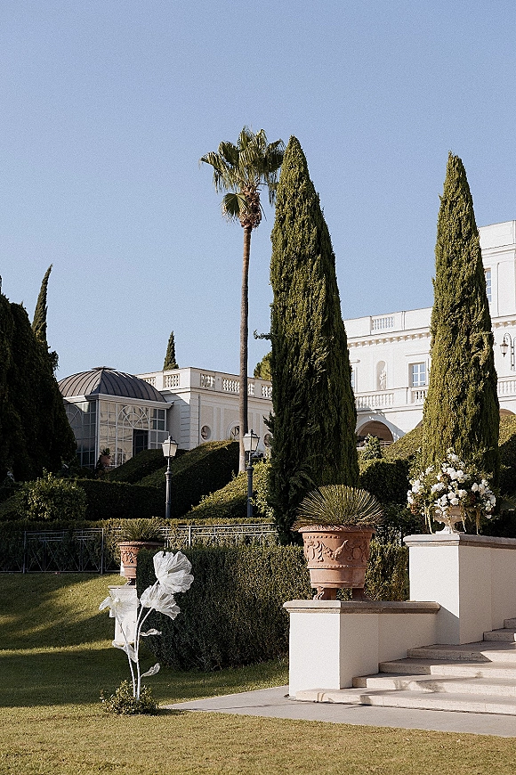 Outdoor wedding venue with wedding venue exterior details like a white estate facade, grand staircase, urn florals, and manicured hedges under blue sky