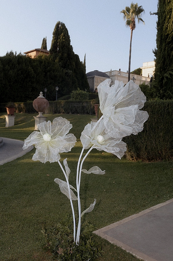 Ceremony aisle decor with oversized wedding flowers in white fabric blooms lining a stone path through manicured estate gardens with hedges and cypress trees