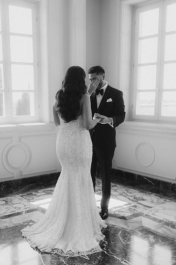 First look moment as the bride in a fitted lace gown approaches the groom in a tuxedo, holding hands by tall windows in soft light