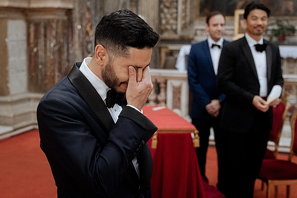 Groom emotional moment as he wipes tears in a black tuxedo at the church altar, groomsmen behind him and a red carpet aisle