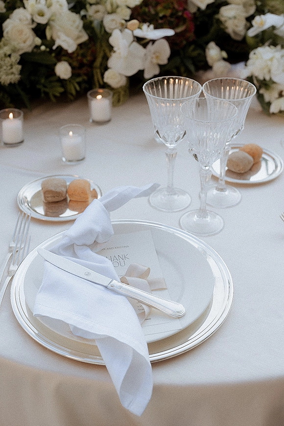 Reception tablescape featuring a wedding place setting with stacked white plates, silver flatware, crystal glasses, tealights, menu card, and floral centerpiece