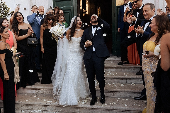 Wedding recessional as bride and groom walk out holding hands, laughing on stone steps while guests toss rose petals by green doors