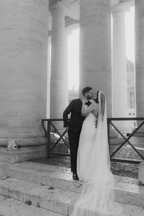 Wedding kiss portrait of bride and groom kissing in a black and white wedding photo on stone steps by a colonnade, cathedral veil trailing