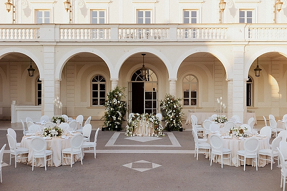 Reception tablescape with outdoor reception tables dressed in white linens, greenery garlands, and tall glass candles in an arched courtyard