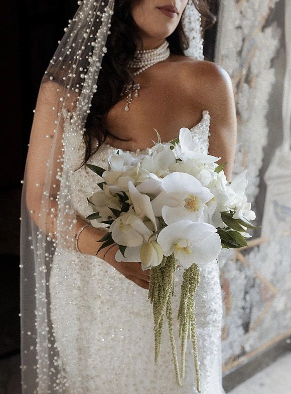 Bridal portrait of a bride holding bouquet of white orchids and greenery, wearing a strapless beaded gown with pearl veil in window light