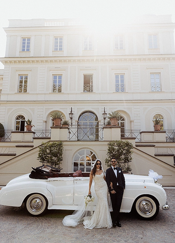 Couple portrait of bride and groom with car, leaning on a classic convertible in sunglasses before a grand staircase facade with potted plants