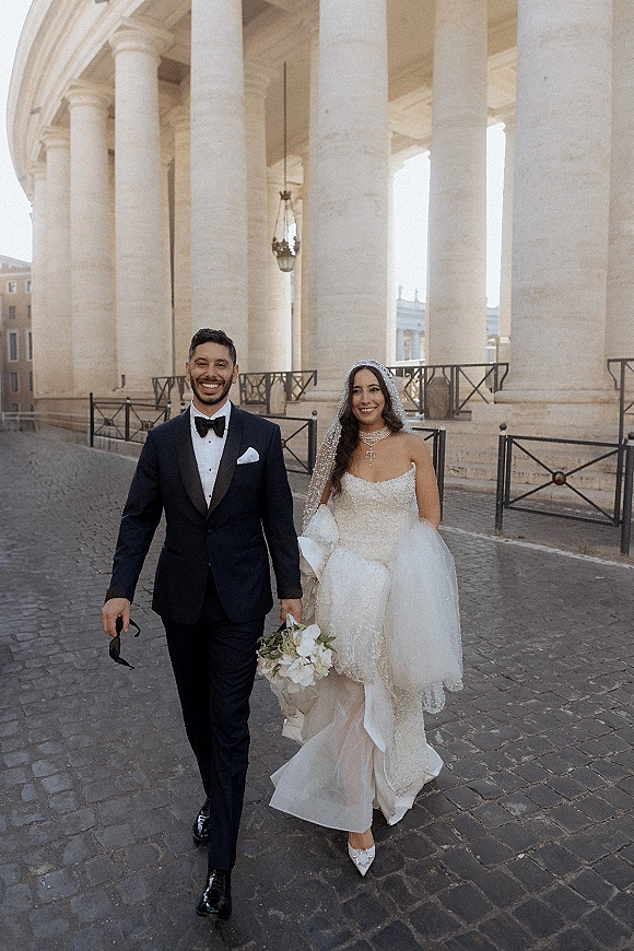 Wedding couple portrait of bride and groom walking arm in arm, she holds a bouquet as stone columns and a lantern line the street