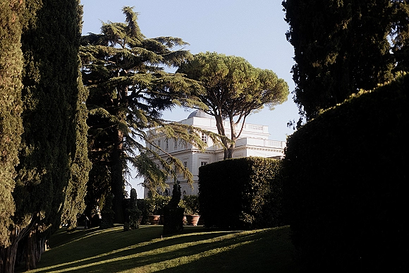 Wedding venue exterior of a white estate wedding venue mansion with manicured hedges, topiary shrubs, and terracotta planters under blue sky