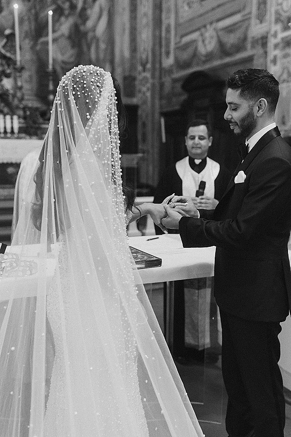 Wedding ring exchange as the groom places a band on the bride’s finger, pearl veil draped behind them at a candlelit church altar