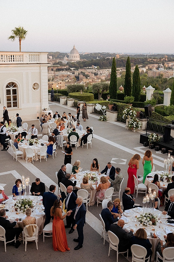 Outdoor wedding reception with round tables and tall glass candle holders, set on a villa terrace with city skyline and dome view