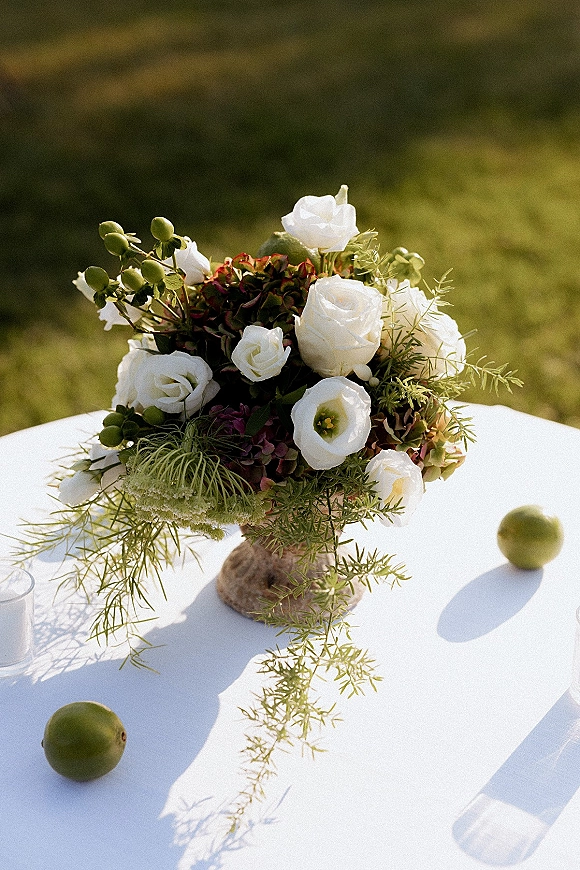 Wedding centerpiece of white roses and greenery in a glass vase with limes on a white tablecloth, set on a sunlit outdoor lawn