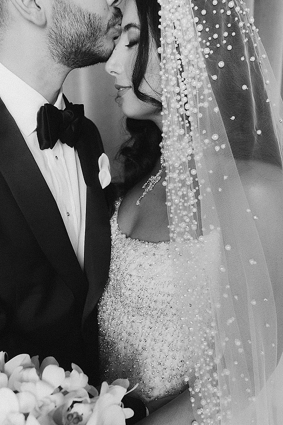Wedding couple portrait of groom kissing the bride’s forehead, her beaded veil and strapless gown framed against an indoor wall backdrop.