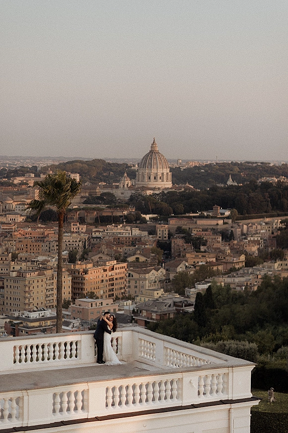 Wedding couple portrait of bride in lace dress and veil kissing groom in black suit on a rooftop terrace with city skyline and dome building behind