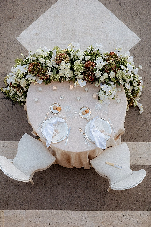 Sweetheart table decor with beige linen, hydrangea and white rose floral garland, and candlelit place settings before a white backdrop