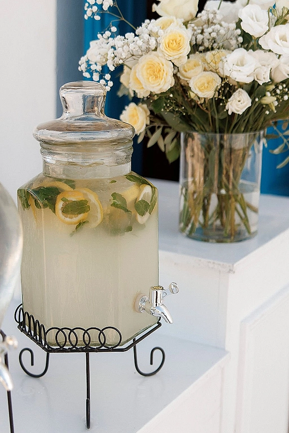 Wedding drink dispenser filled with lemon mint infused water on a black metal stand, garnished with slices, beside white roses on a table with blue drapery