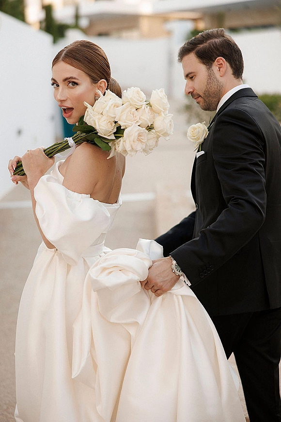 Couple portrait of bride holding bouquet of white roses beside groom in tuxedo on an outdoor walkway with white walls and greenery
